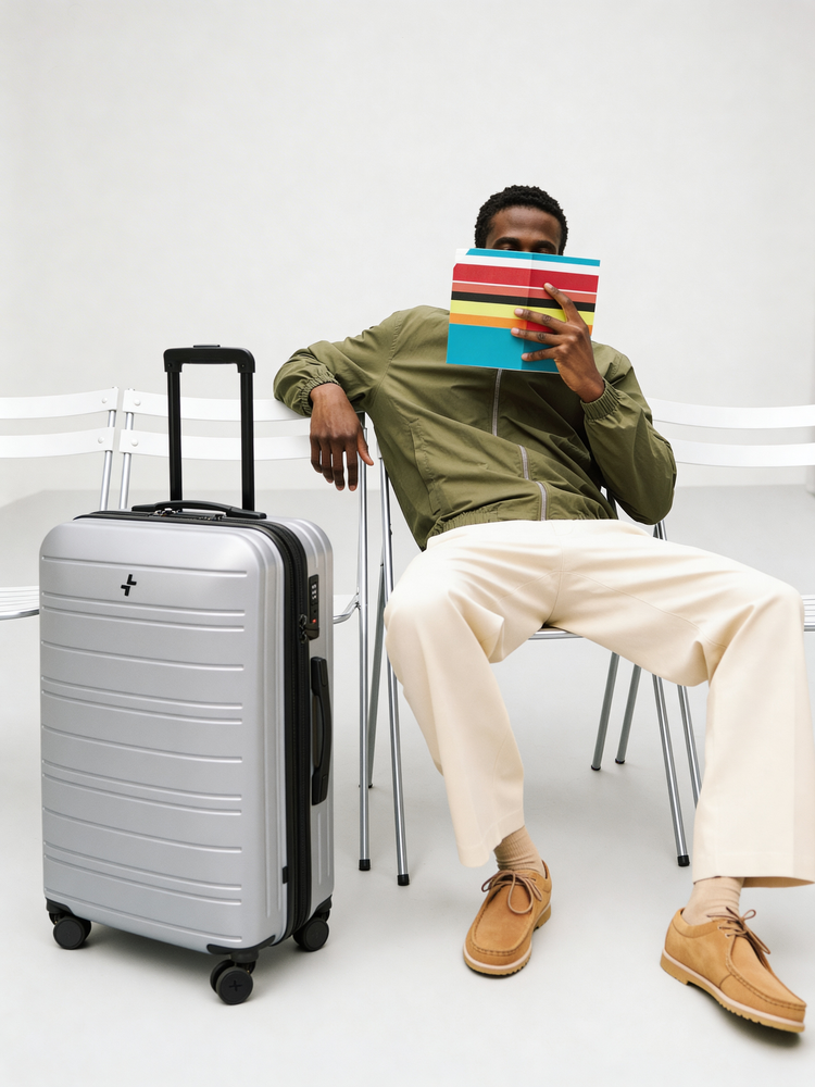 Person sitting with a colorful book, holding it over their face, next to a silver Bentley suitcase on a white background.